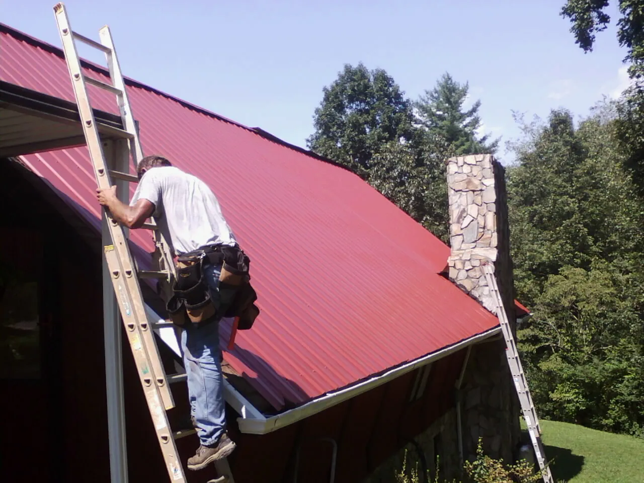 Roofer on a ladder beside a red metal roof and stone chimney — Soffit & Fascia Repair by Roofing Brothers in Newfield